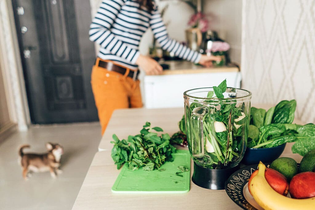 woman making a smoothie with spinach while her dog hangs out in the kitchen with her